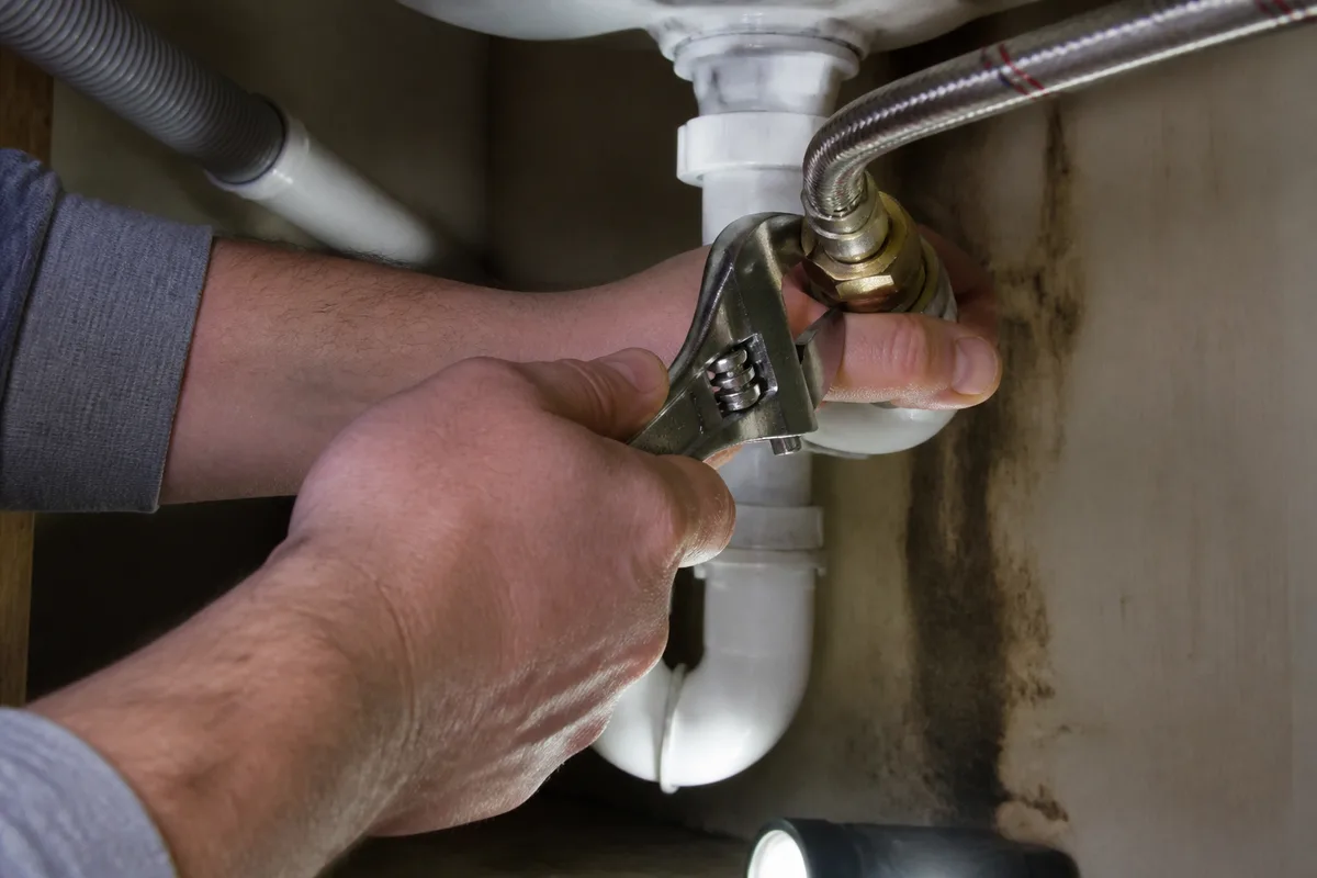 Mains d’un plombier resserrant un raccord de canalisation sous un évier avec une clé